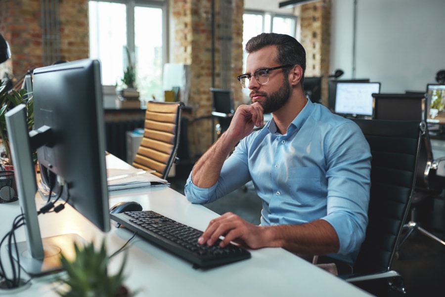 Side view of young bearded man in eyeglasses and formal wear working on computer